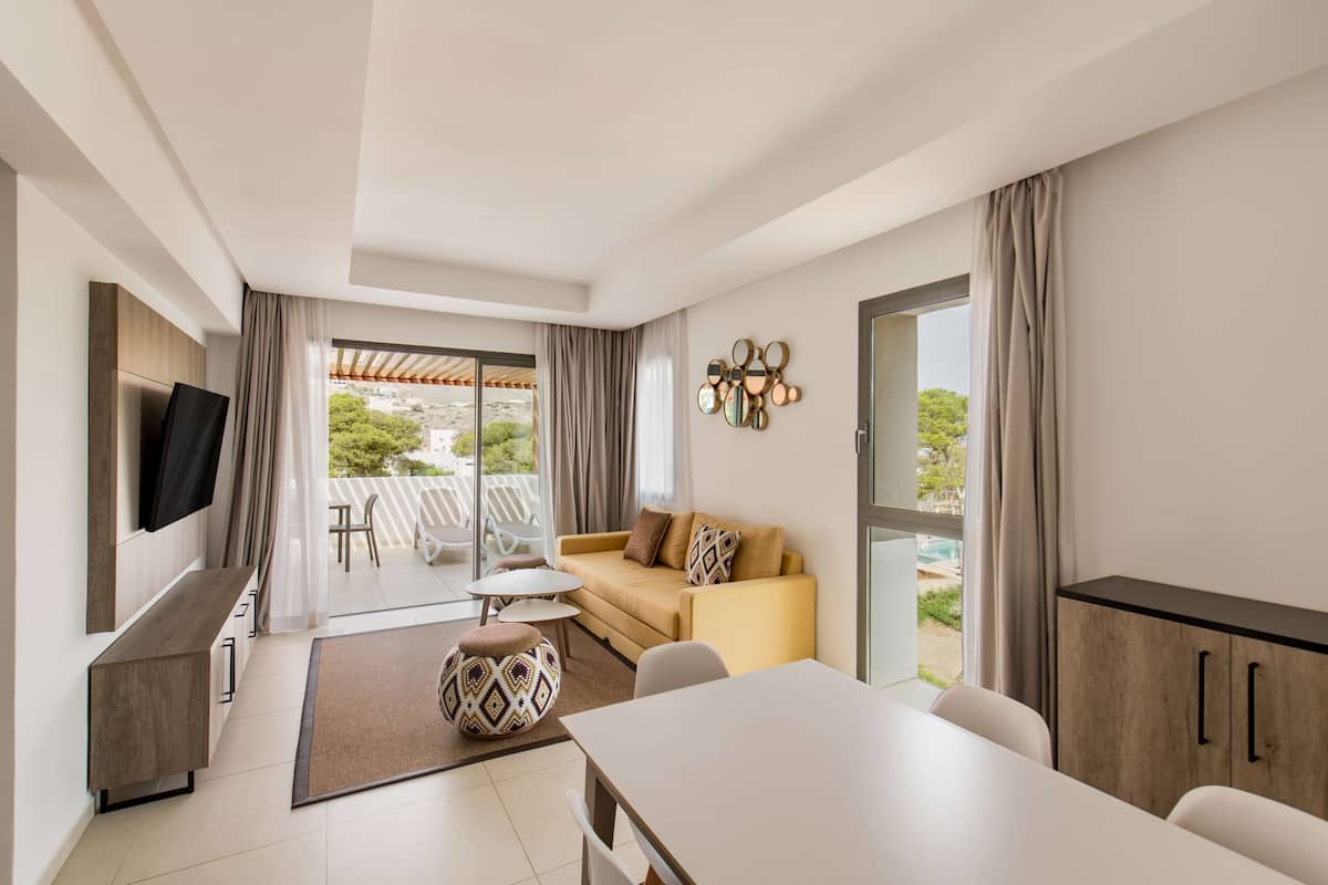 A modern living room with a sofa, a TV, a dining table, and a view of the outdoors.