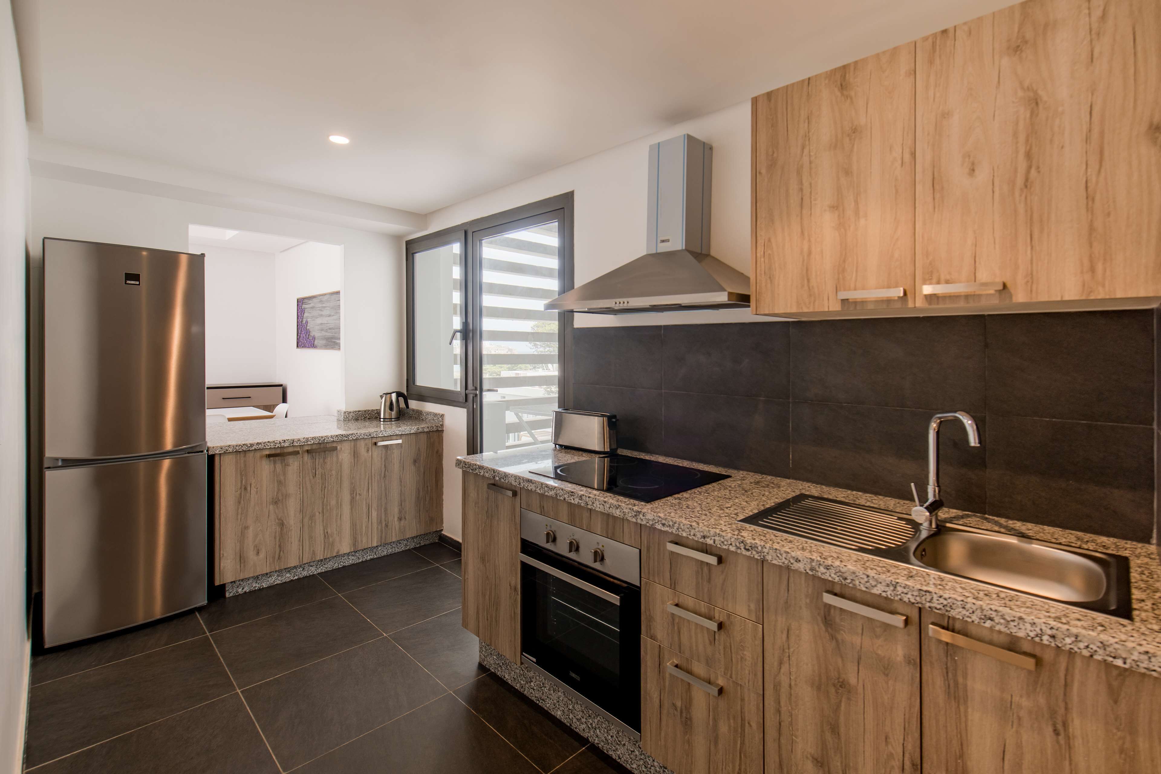 A modern kitchen with wooden cabinets, stainless steel appliances, a built-in oven, and a large window with blinds.