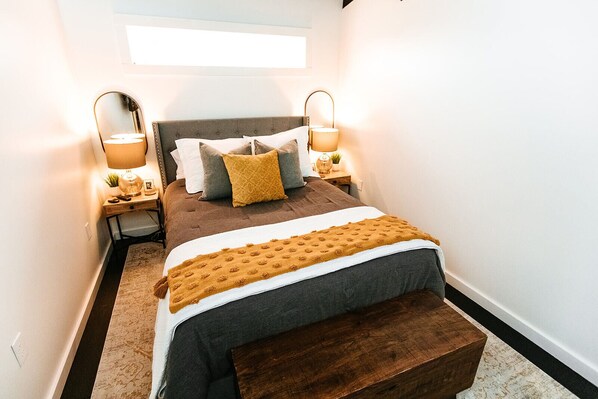 A narrow hotel room with a bed, two bedside lamps, a small table with a plant, and a bench at the foot of the bed.