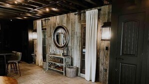 A rustic interior with a wooden floor, a round mirror, a dark door, and a bar area with stools.