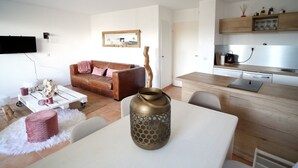 Ein modernes Wohnzimmer mit einem braunen Ledersofa, einem weißen Couchtisch und einem Esstisch mit einer dekorativen Vase. Im Hintergrund ist eine Küche mit Holzschränken und einem Einbauofen zu sehen.