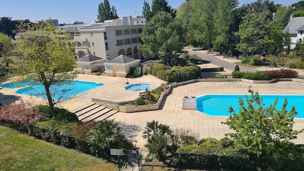 Ein Poolbereich mit mehreren Schwimmbecken, umgeben von viel Grün und einem Gebäude im Hintergrund.