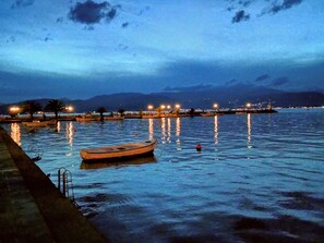 A calm waterfront at dusk with boats anchored near the shore, a pier extending into the water, and distant city lights. - Cosy Villa With Garden, Near the Beach in Greece (Argos-Mykines)