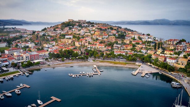 Une ville côtière avec une colline centrale où se trouvent des bâtiments historiques, un port de plaisance avec des bateaux et une vue dégagée sur l’eau et les collines environnantes.