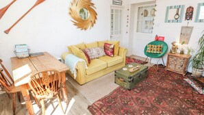 A cozy living room with a yellow sofa, wooden dining table, and chairs. A green armchair and a patterned rug are also visible. - The Cottage (Penzance)
