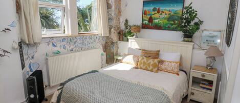 Quarto com cama, radiador, janela com cortinas, quadro na parede, mesa de cabeceira com abajur e planta.