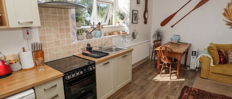 Een keuken met een fornuis, een spoelbak en een houten aanrecht. Er is een eettafel met stoelen en een gele bank in de woonkamer.