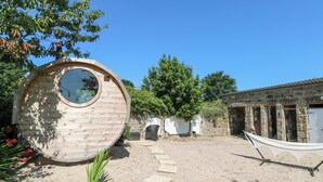 A wooden spherical structure with a circular window, surrounded by a gravel path, a hammock, and a stone building in the background. - The Cottage (Penzance)