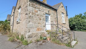 A stone building with a white door and windows, a small porch with steps, and potted plants. - The Cottage (Penzance)