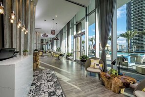 A modern hotel lobby with a long reception desk, seating area, and large windows offering views of a pool and palm trees.