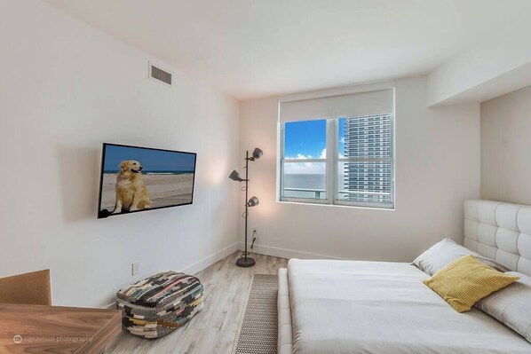 A bedroom with a bed, a window with blinds, a floor lamp, a rug, a television on the wall, and a small ottoman. - Opulent Three Bedroom Condo Panoramic Ocean Views (Hollywood)
