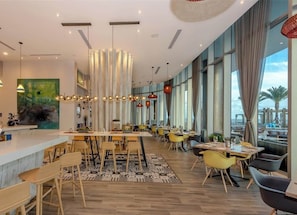 Un ristorante moderno con un lungo bancone, diversi tavoli e sedie. Grandi finestre offrono vista all'esterno.