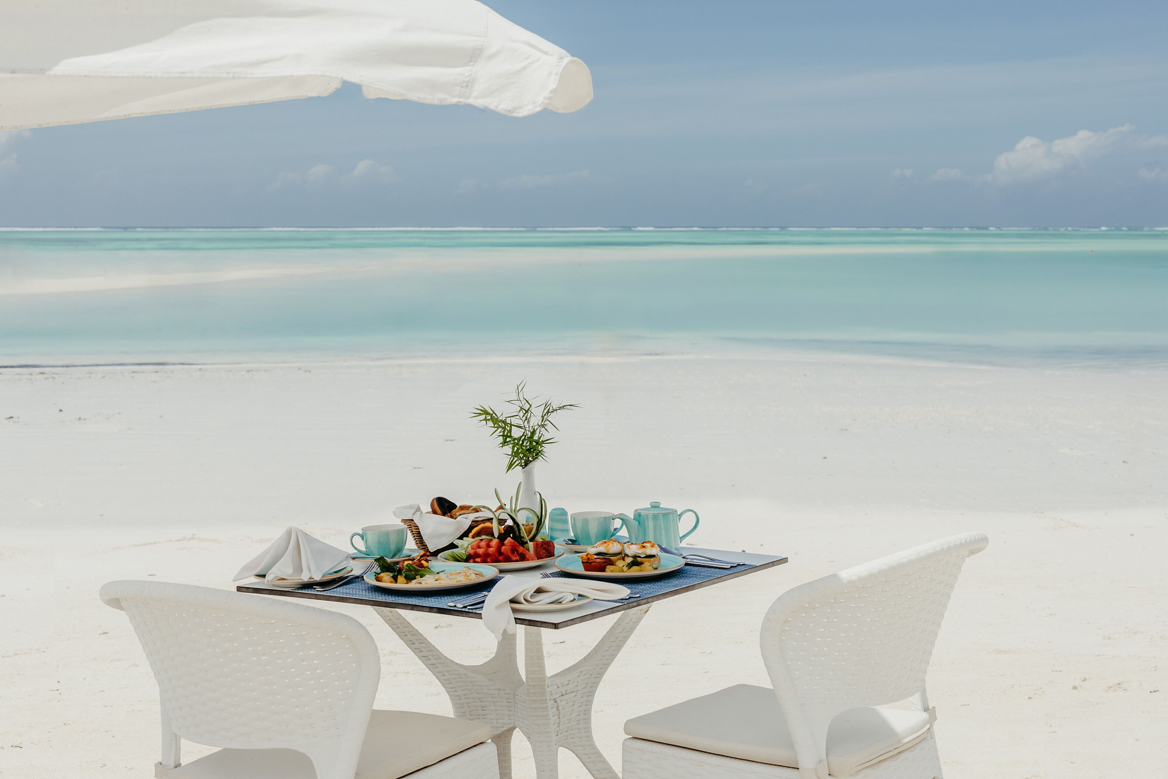 A dining table with plates of food, a vase with a plant, and a teapot on a beach with a clear view of the ocean.