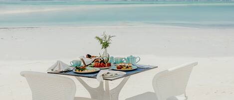A dining table with plates of food, a vase with a plant, and a teapot on a beach with a clear view of the ocean.