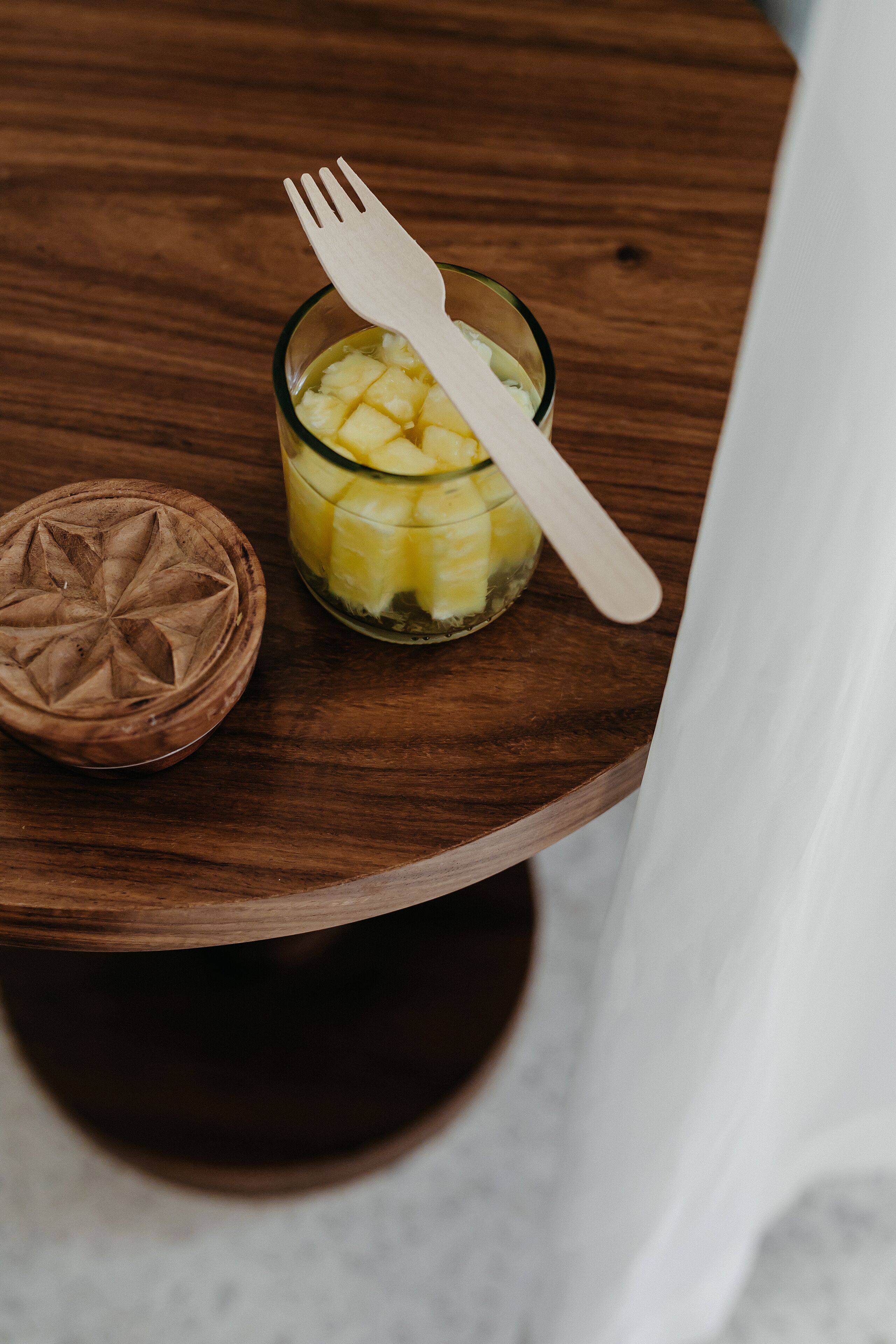 A wooden table with a glass containing pineapple chunks and a wooden fork, and a wooden container with a star design.