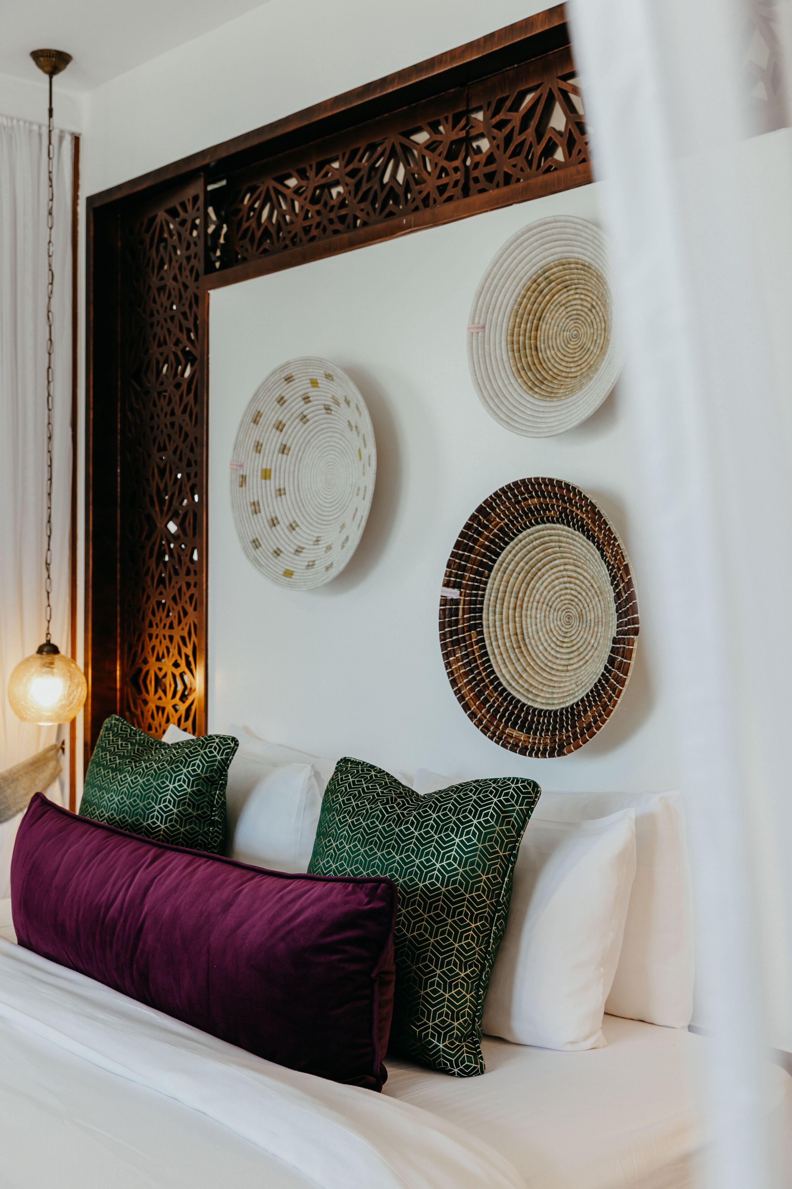 A neatly arranged bed with patterned pillows, a decorative wall panel, and two woven wall hangings.