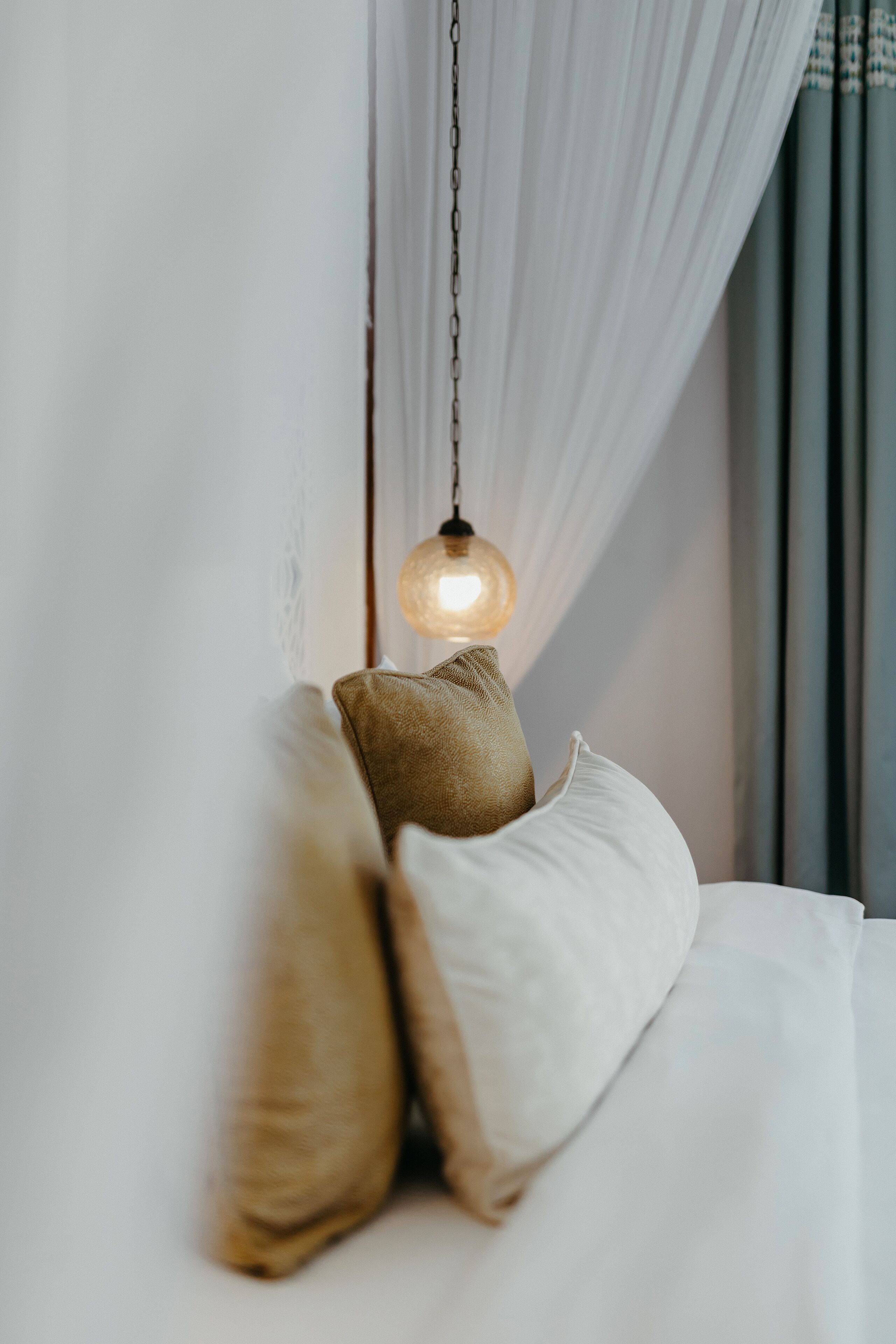 Een bed met witte beddengoed en twee kussens, een hanglamp en dunne gordijnen.