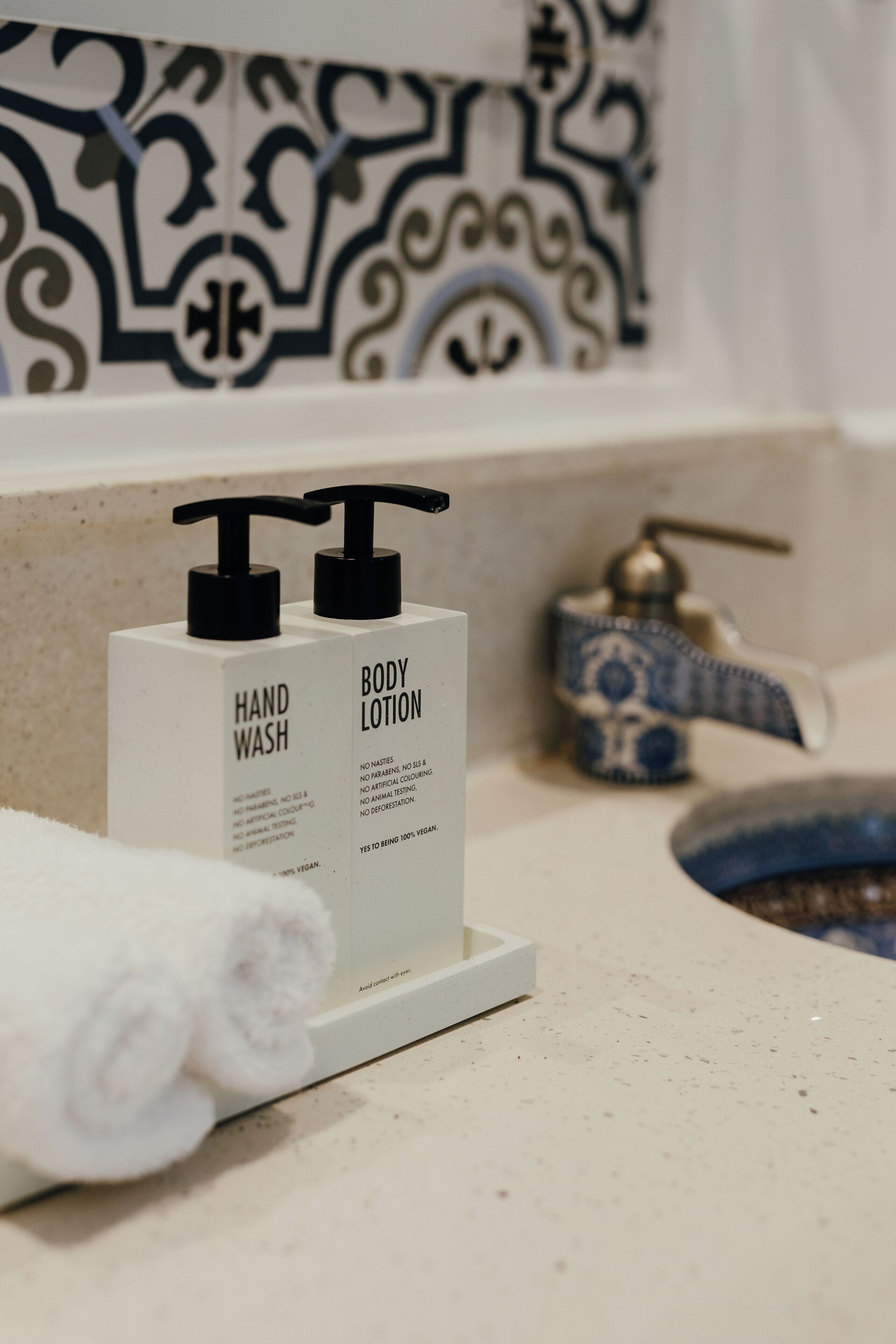 A bathroom with a white countertop, two bottles of hand wash and body lotion, a blue and white patterned faucet, and a white towel rolled up.
