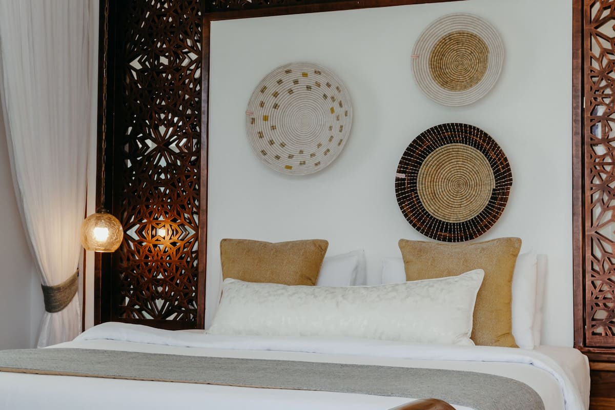 A bedroom with a wooden headboard, a bed with white and beige bedding, decorative wall art, and a wooden partition with intricate cut-out patterns.