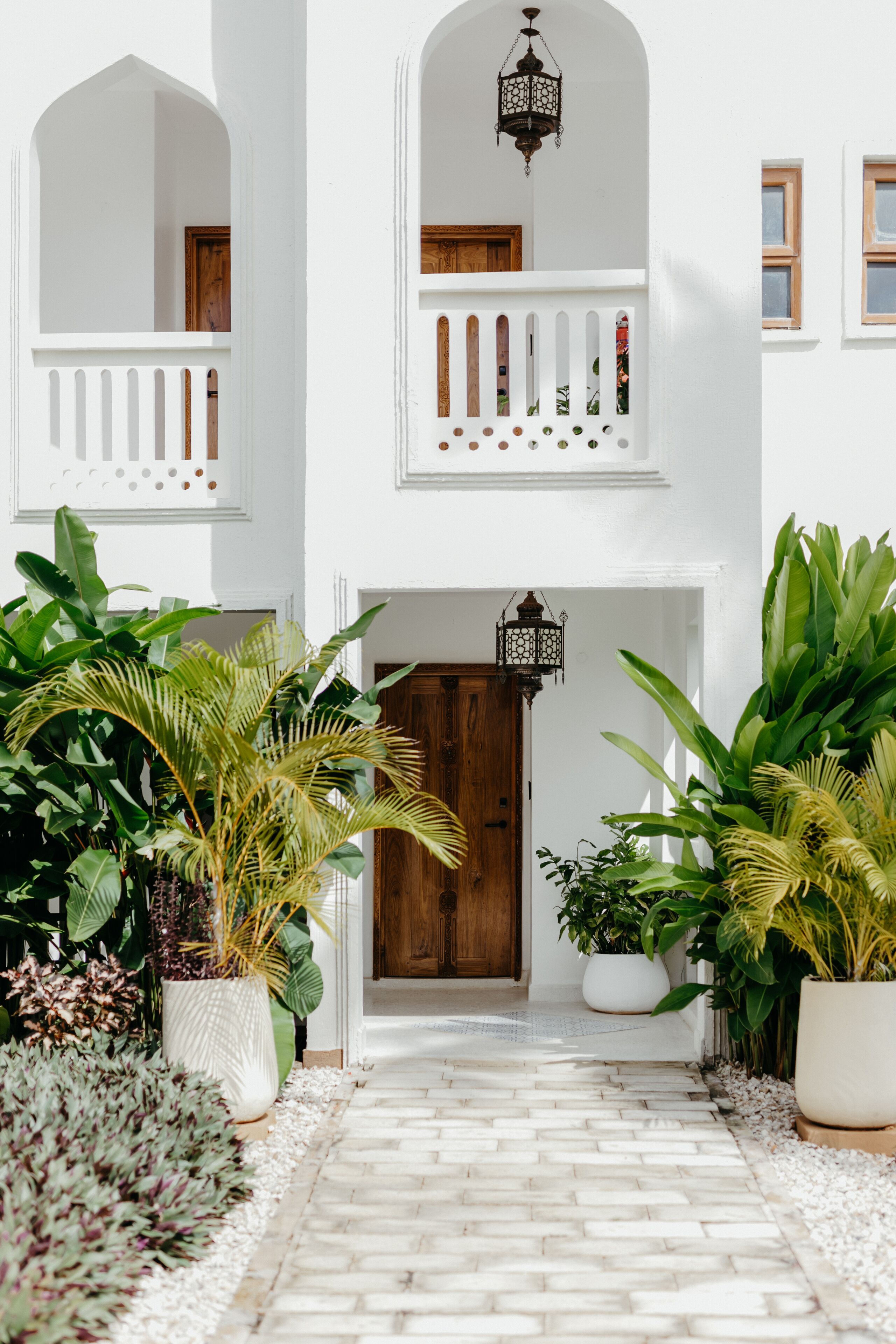 A white building with a central wooden door, potted plants, and a tiled pathway leading to the entrance.