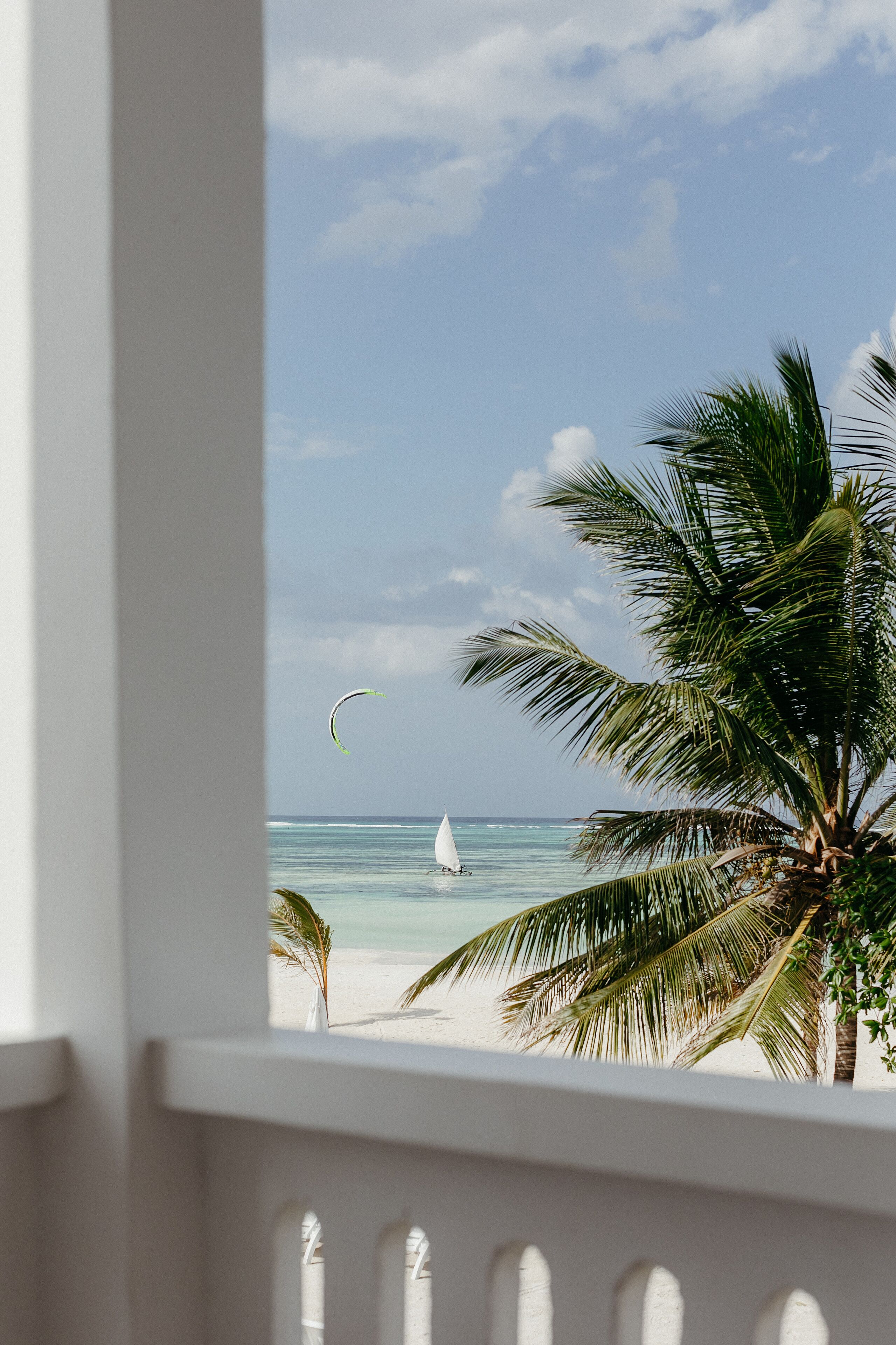 A beach view from a balcony with a sailboat and palm trees.
