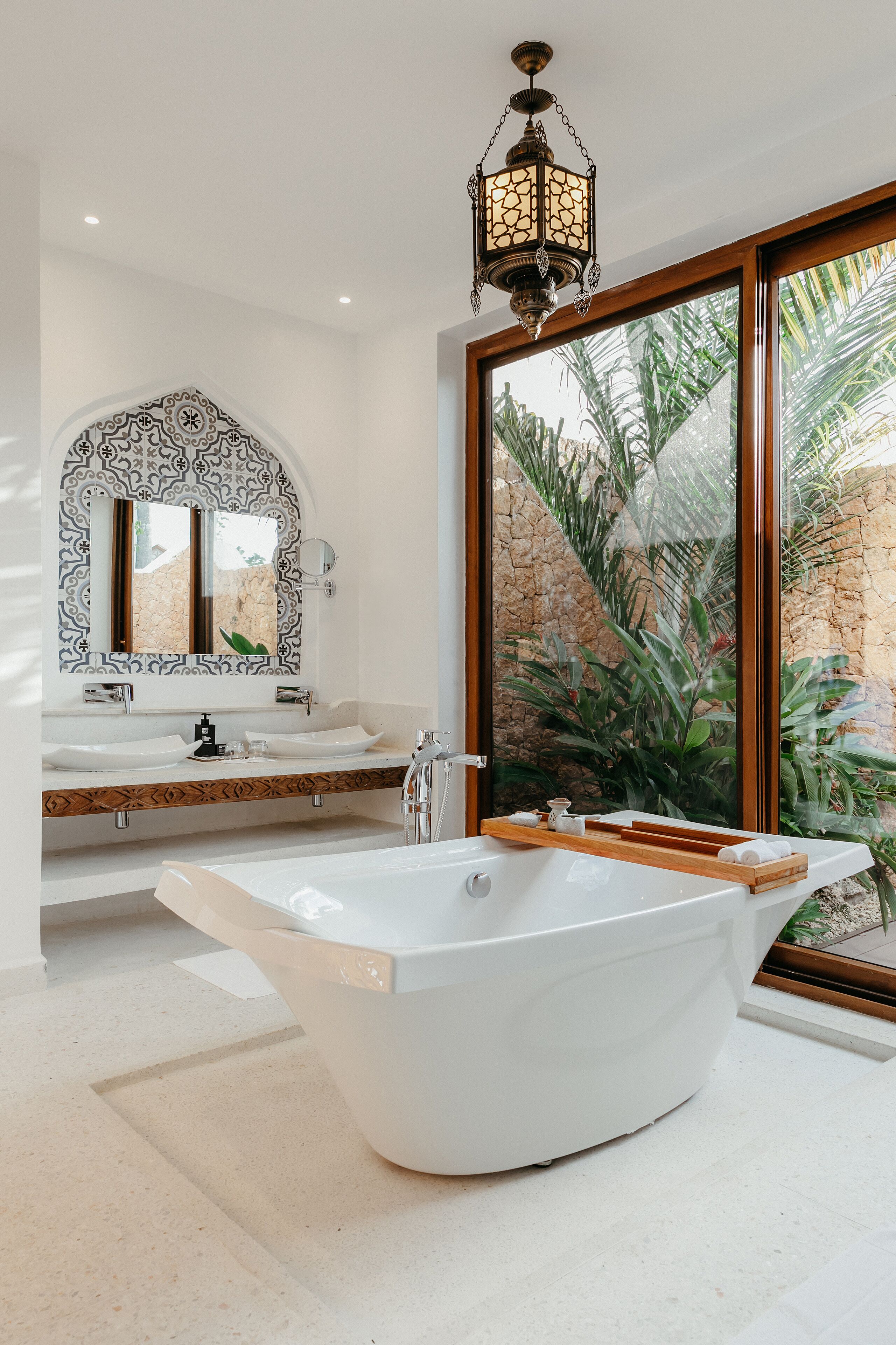A modern bathroom with a freestanding bathtub, a large mirror with a decorative frame, and a view of a garden through a large window.