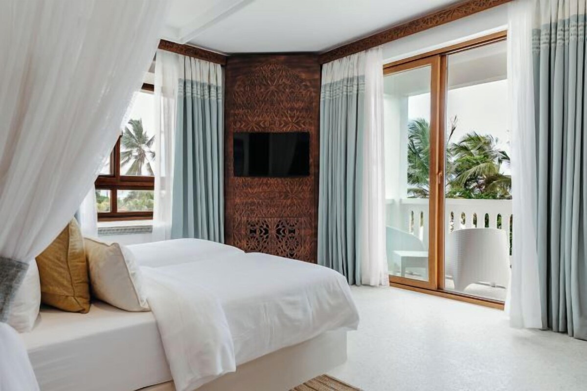 A bedroom with a bed, a TV mounted on the wall, and a balcony with a view of palm trees.
