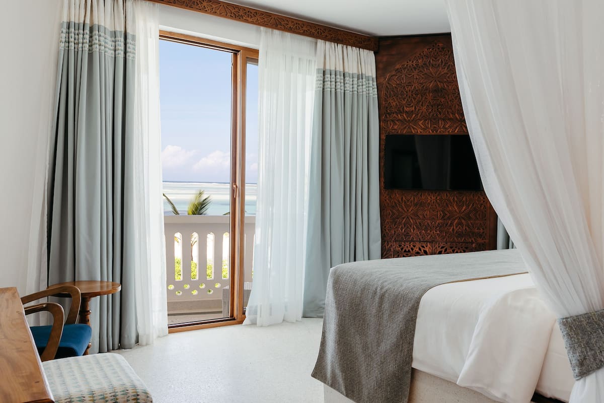 A hotel room with a bed, a chair, a TV, and a balcony with a view of the ocean.