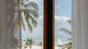 A room with a large window overlooking a beach with palm trees and a sailboat. - LUX Marijani Zanzibar (Pwani Mchangani)