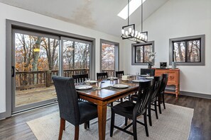A dining room with a large wooden table set for six, surrounded by black chairs. There are large windows providing a view of a wooden deck and trees. A modern chandelier hangs above the table, and a wooden cabinet is against the wall. - Gorgeous, spacious 4 bedroom home in Wintergreen Resort (Roseland)