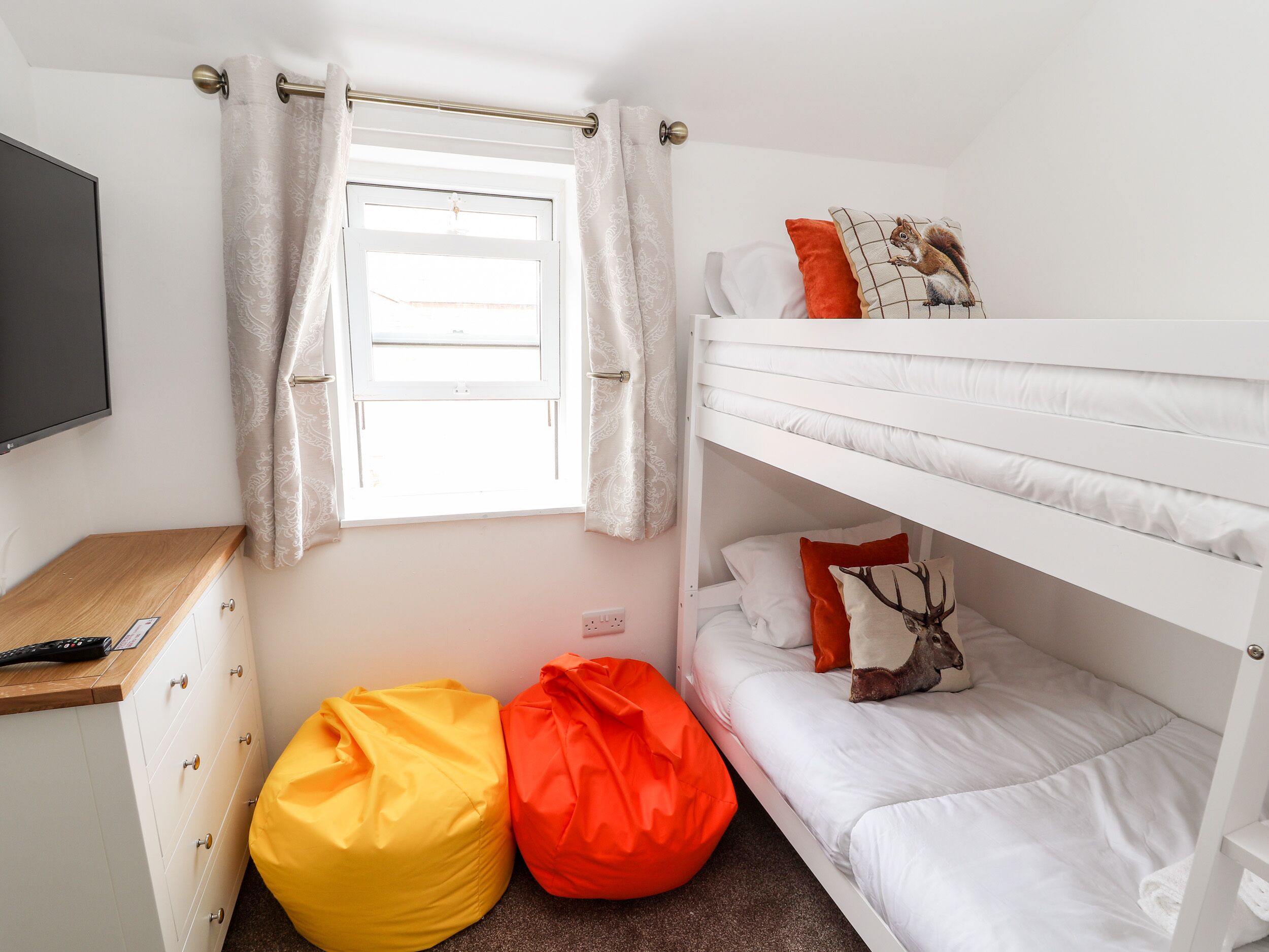 Uma cama beliche com roupa de cama branca, uma janela com cortinas, uma escrivaninha com um controle remoto e dois sacos de feijão.