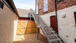 Un estrecho callejón con una escalera de metal que lleva a una puerta de madera, junto a un edificio de ladrillo con una pared blanca y una puerta roja.