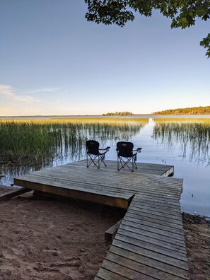 Une jetée en bois avec deux chaises, donnant sur un lac paisible, entourée de hautes herbes et d’arbres.
