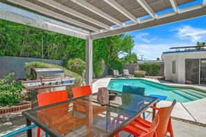 A covered outdoor area with a barbecue grill, a glass table, and orange chairs.