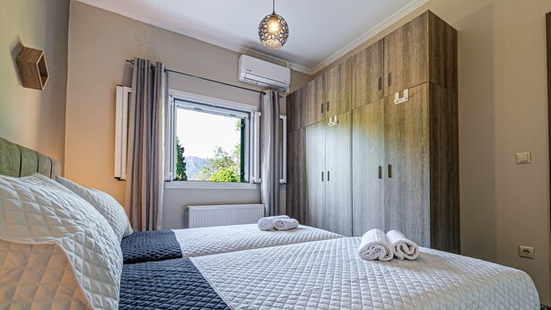 A hotel room with two beds, a large wardrobe, a window with curtains, and an air conditioning unit. - Thomas House (Corfu)
