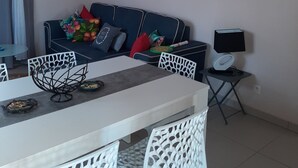 Ein modernes Wohnzimmer mit Esstisch in Weiß, weißen Stühlen, einem blauen Sofa mit bunten Kissen, einem Gemälde einer Wasserfall und einem kleinen Beistelltisch mit Lampe.