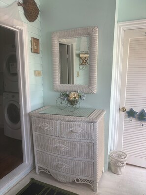 A room with a white wicker dresser, a mirror with a woven frame, a vase with flowers, and a door with a decorative handle.