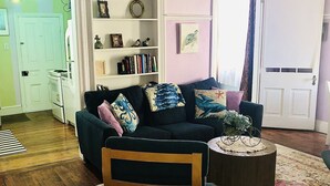 A cozy living room with a dark blue sofa, a wooden chair, a small round table, and a bookshelf filled with books and decorative items.