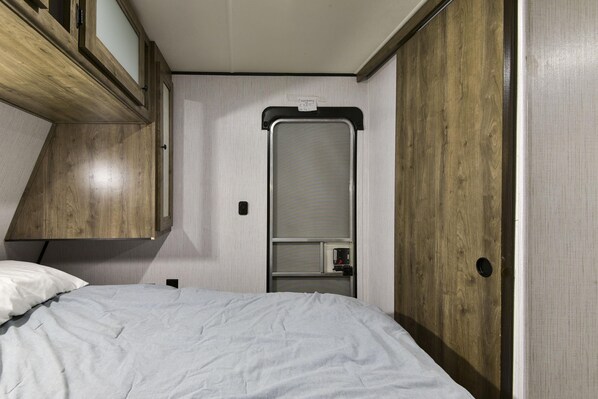 A compact bedroom with a bed, a wooden wardrobe, and a small window. - Givens Glamper Heartland W 3 Beds, Outdoor Pool, Views for Days (Branson)