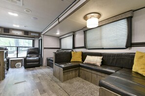 Un interior de caravana acogedor, con un sofá modular, un sillón reclinable y una ventana con persianas.