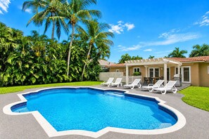 A swimming pool with a unique shape, surrounded by palm trees and a covered patio area with lounge chairs.