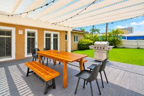 Une terrasse extérieure avec une table et des bancs en bois, un barbecue et un espace abrité agrémenté de guirlandes lumineuses.