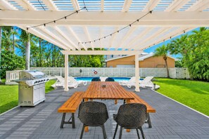 An outdoor patio with a wooden table and chairs, a barbecue grill, and a pool in the background.