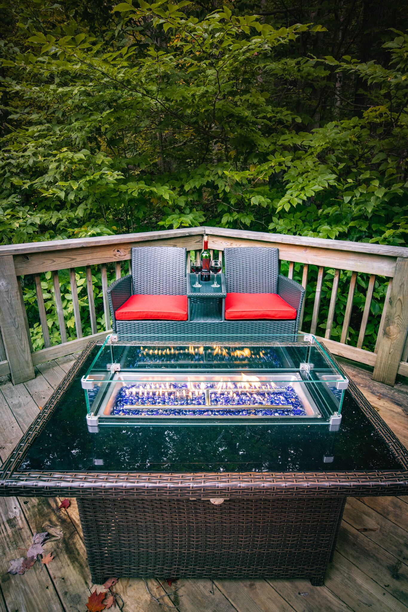 A glass-top table with a chess set, surrounded by wicker chairs with red cushions, on a wooden deck with a wooden railing.