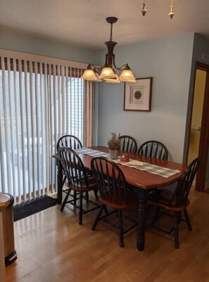 A dining area with a wooden table, six chairs, a chandelier, and a window with blinds.