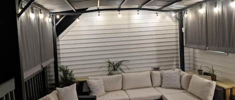 Un espacio al aire libre techado con sofá y sillones, una mesa y plantas en macetas.