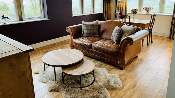 Un salon chaleureux avec un canapé en cuir, une table basse ronde en bois et une table à manger avec des chaises. De grandes fenêtres offrent une vue sur l’extérieur.