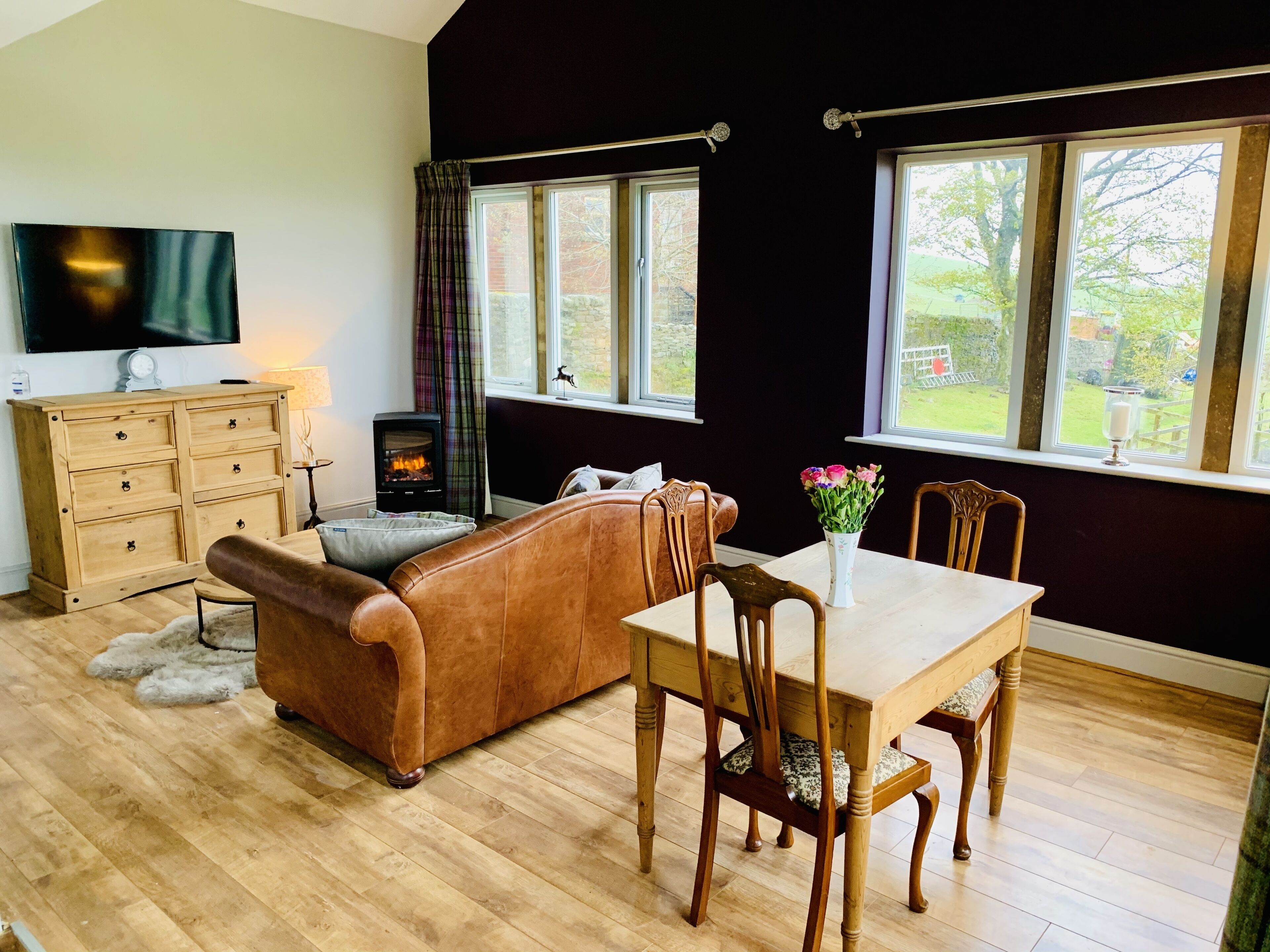 A cozy living room with a brown leather sofa, wooden dining table, and chairs. A television is mounted on the wall, and a fireplace is visible in the corner.