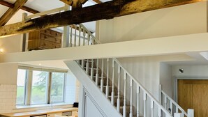 Un espace de vie de style loft avec des poutres apparentes, un escalier blanc, un coin cuisine et un canapé en cuir marron.