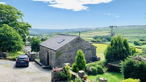 Une charmante maison en pierre avec un toit en pente, une voiture garée devant, et une vue sur des collines et des champs vallonnés.